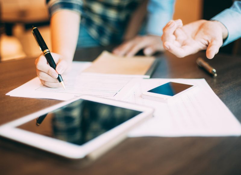 Two businesspeople standing at table in office, woman filling in paper with pen, man explaining something to her. Digital tablet and mobile phone lying on table